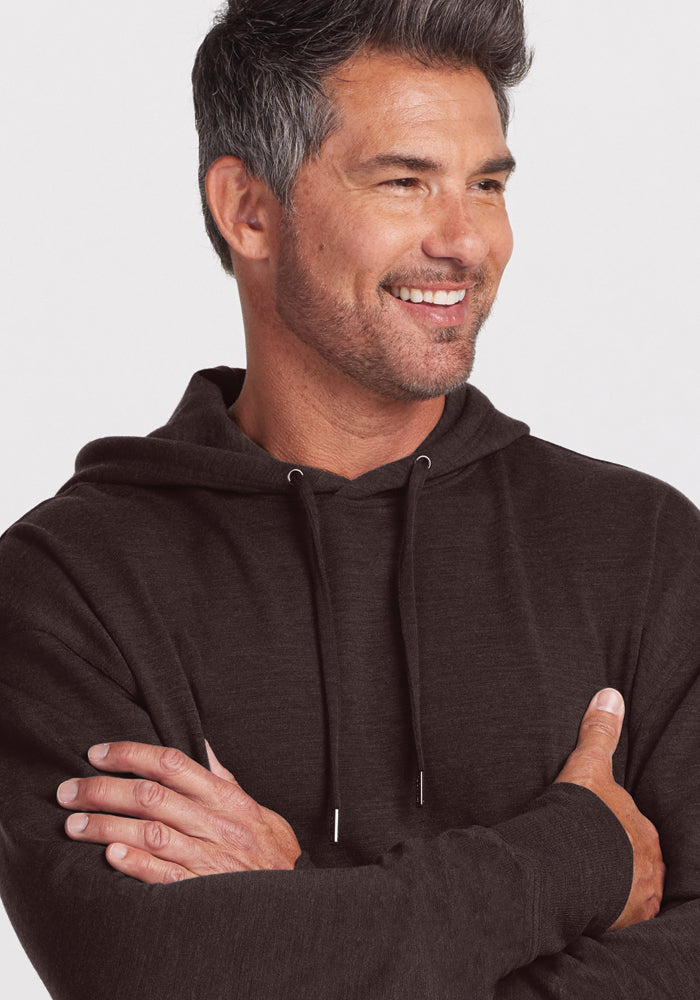 A man with short, dark hair and a beard is smiling and looking to his left. He is wearing a Maple Melange Chase Hoodie from Woolx made of soft Merino wool and has his arms crossed. The background is plain white.