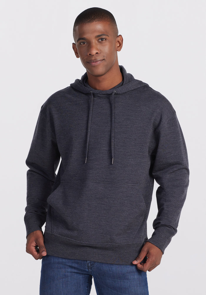 A person stands against a plain background, wearing the Woolx Chase Hoodie in Pebble Grey Melange with drawstrings and blue jeans. The individual is looking at the camera with a slight smile, their hands relaxed at their sides with thumbs in their pockets.