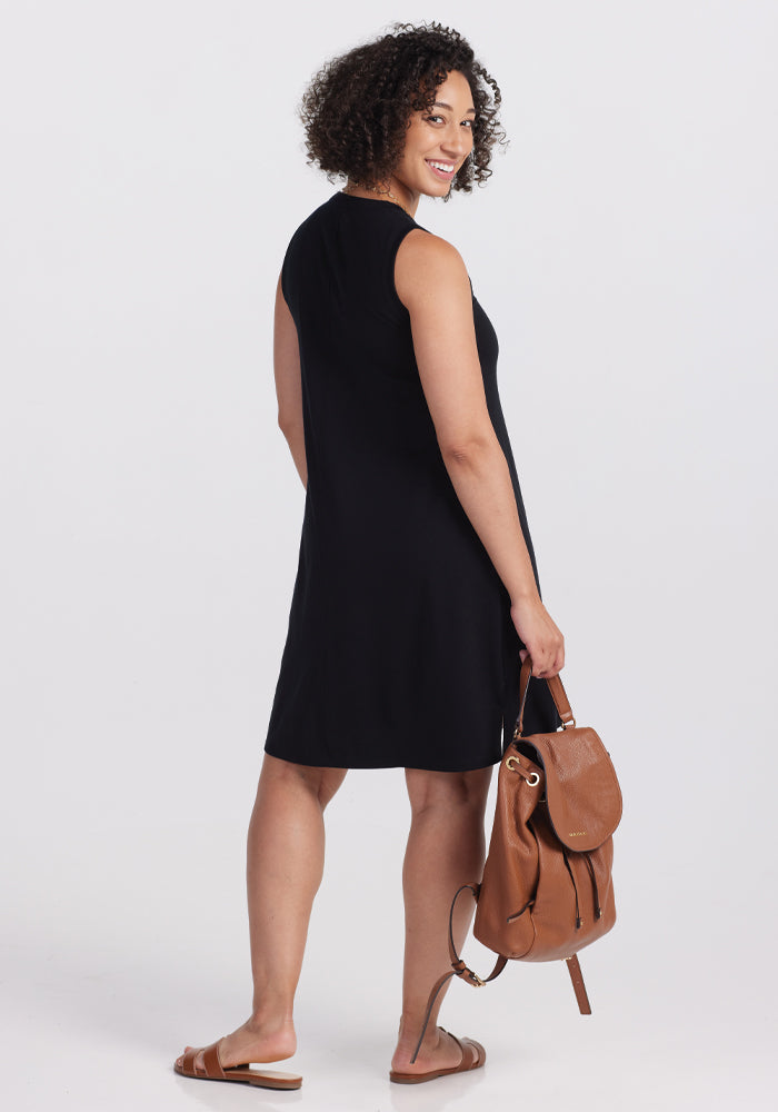 A curly-haired person smiles over their shoulder while standing, wearing a sleeveless Woolx Clara Dress in black with sandals and holding a brown backpack. The plain white background subtly highlights their stylish ensemble.