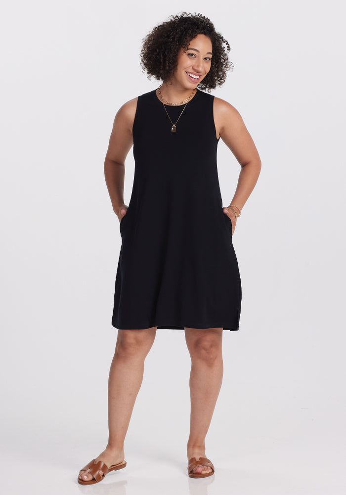 A person with curly hair smiles in a stylish Woolx Clara Dress - Black, paired with brown sandals. They have their hands in their pockets, standing against a plain white background. 