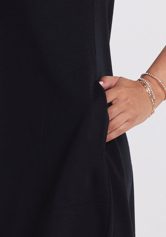 A person in a chic Woolx Clara Dress - Black with their hand in a pocket. Several gold and silver bracelets adorn their wrist, adding elegance. The plain white background highlights the stylish ensemble.