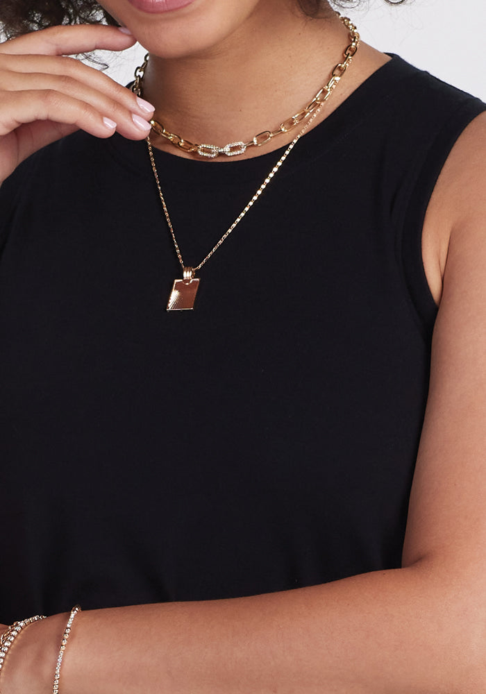 A person is elegantly styled in a Clara Dress - Black by Woolx, adorned with two gold necklaces: one featuring a textured chain and the other a rectangular pendant, paired with a delicate gold bracelet.