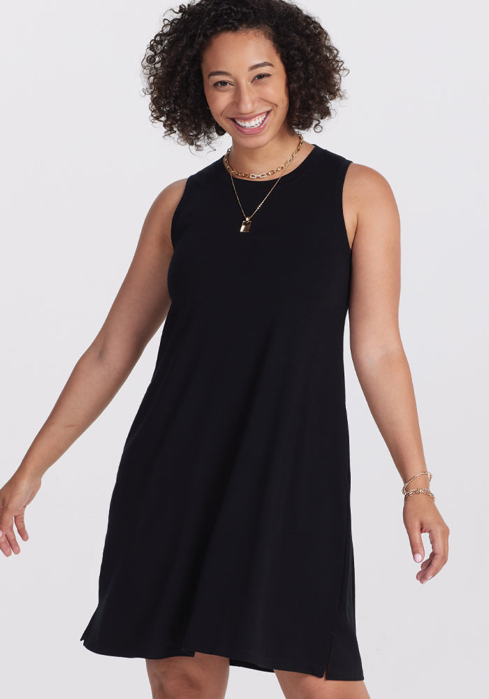 A person with curly hair is smiling while wearing a Black Clara Dress by Woolx, accented with gold jewelry, standing against a plain white background.