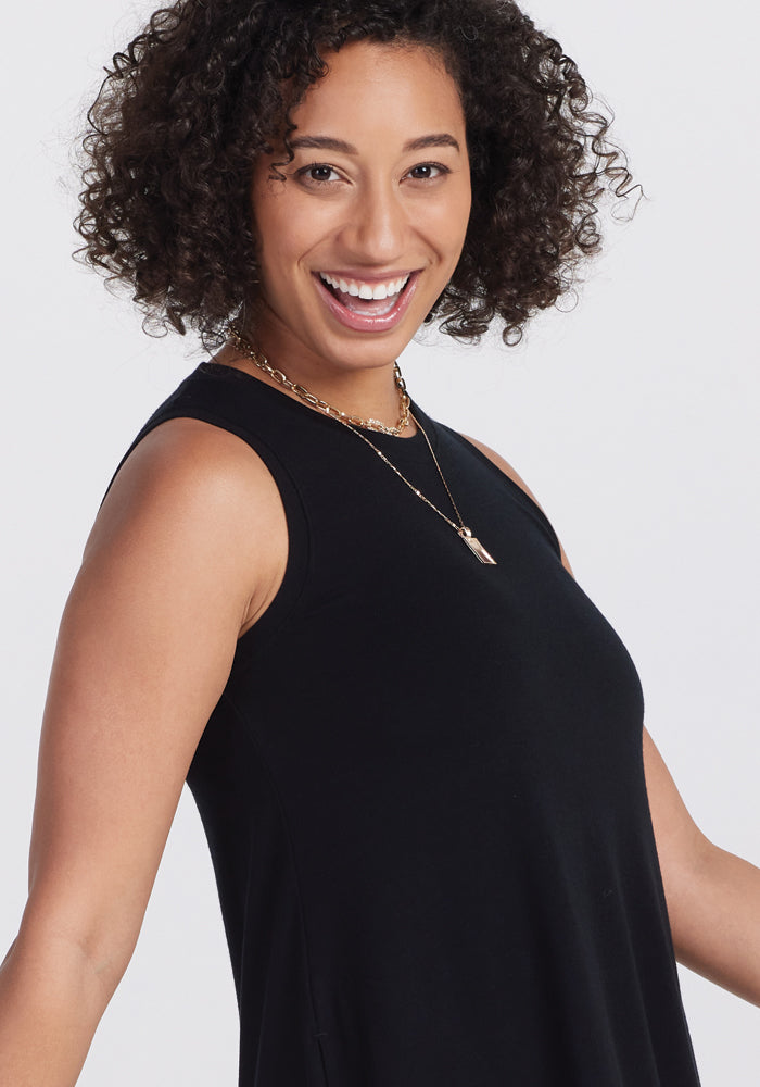 A person with curly hair smiles elegantly in a Clara Dress - Black by Woolx, complemented by a gold chain necklace with two pendants, against a simple white background.