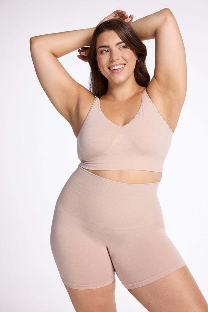 A smiling woman with long brown hair is posing with her arms raised behind her head. She is wearing a matching set of Woolx’s Marley Shapewear Shorts and a high-waisted sports bra, both in light beige and made from merino wool for ultimate comfort. The background is plain white. 