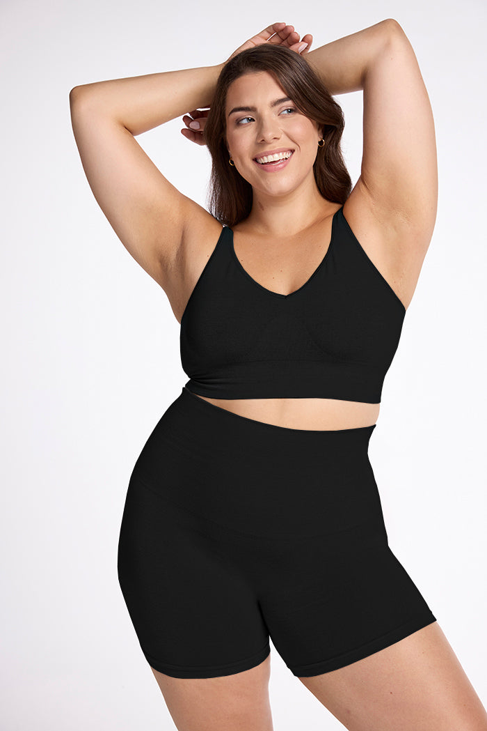 A person with long hair is smiling and posing with their arms lifted behind their head. They are wearing a black sports bra and the Marley Shapewear Shorts from Woolx, and the background is plain white. 