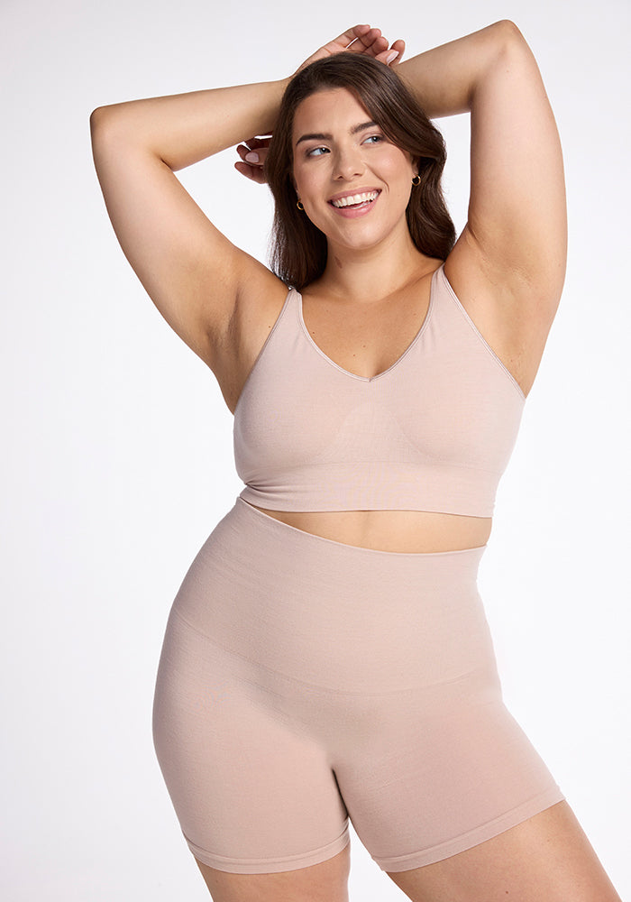 Wearing the Marley Shapewear Shorts in Sand by Woolx, a woman smiles confidently as she highlights the gentle compression of her outfit. One arm is raised while her other hand rests on her head against a plain white background. 