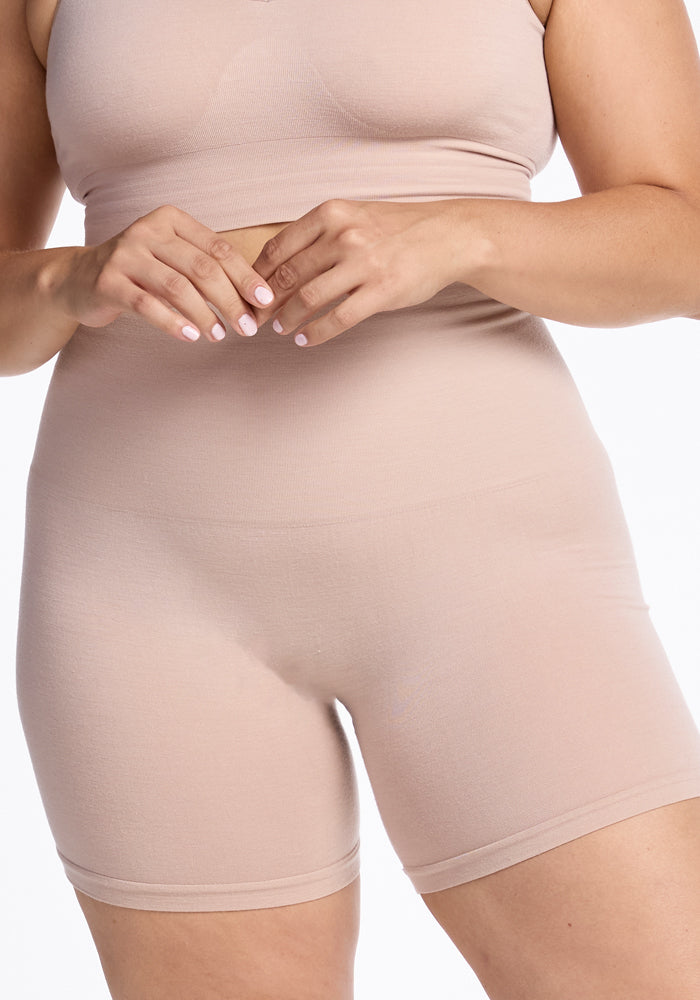 A person dressed in a beige seamless matching set consisting of the Woolx Marley Shapewear Shorts and a crop top made from merino wool. The person's hands, adorned with light pink nail polish, are positioned in front of their torso. The background is plain white.