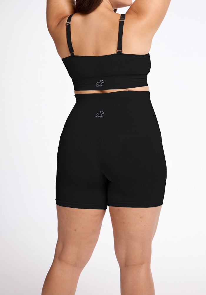 A person facing away, wearing a black sports bra and Marley Shapewear Shorts from Woolx, featuring high-waisted, seamless construction. The outfit displays small logo designs on the back, set against a plain white background.