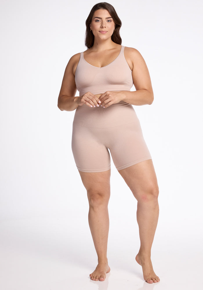 A woman stands barefoot against a plain white background, wearing the nude-colored seamless tank top and Woolx's Marley Shapewear Shorts. She has a slight smile on her face and her hands are gently clasped in front of her.