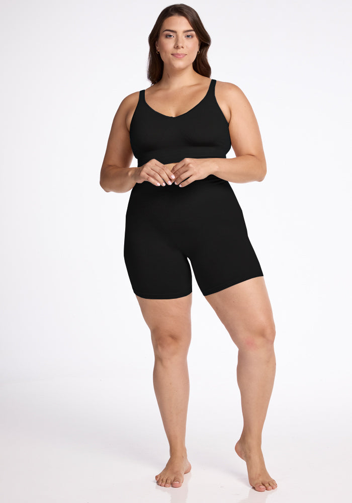 A woman stands against a plain white background, wearing Woolx's Marley Shapewear Shorts, a black full-coverage body shaper that resembles a tank top and shorts. Featuring seamless construction, she rests her hands gently together near her waist and has a confident, relaxed expression. She is barefoot.