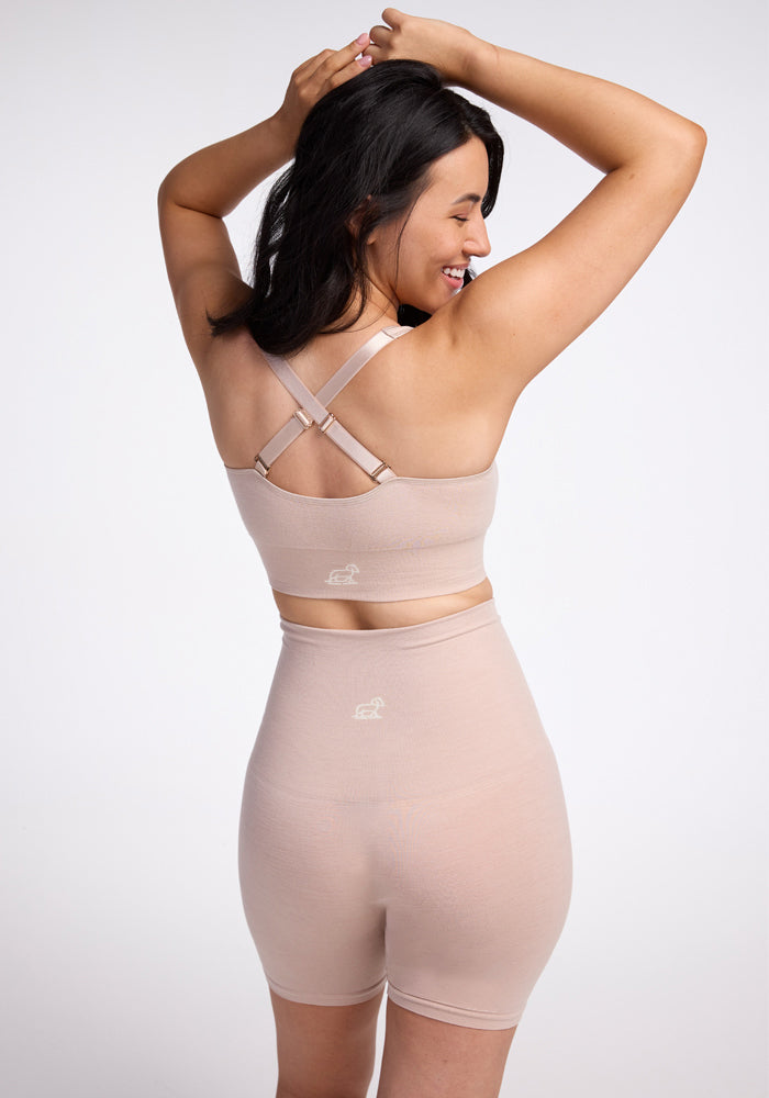 A woman with dark hair, her back facing the camera, is wearing a seamless Marley Shapewear Shorts and sports bra set from Woolx. The beige outfit ensures comfort, and she has her arms raised in a smile. A small logo decorates both the bra and shorts, all set against a plain white background.