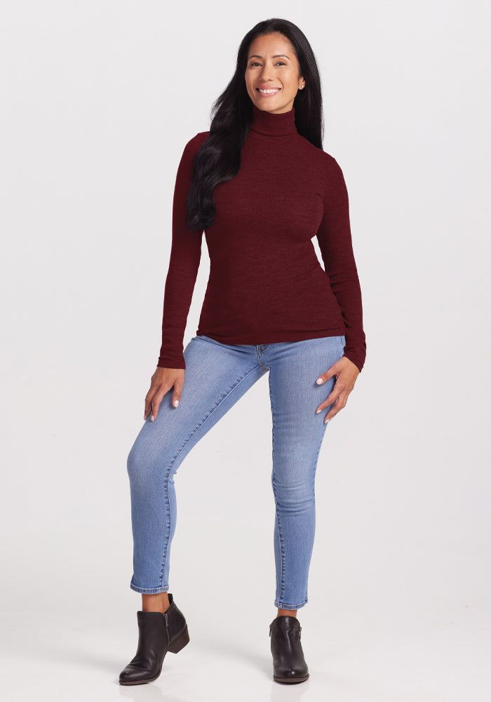 A woman in a Woolx Sage Ribbed Turtleneck - Cranberry Melange pairs it confidently with light blue jeans. She stands with one leg slightly bent, wearing brown ankle boots against a plain white background.