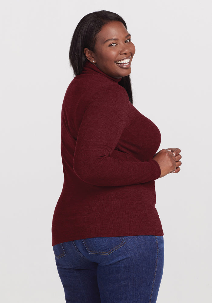 A smiling person with long hair, dressed in a Woolx Sage Ribbed Turtleneck in Cranberry Melange and blue jeans, stands sideways while looking back at the camera.