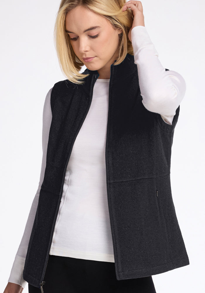 A woman with blonde hair is wearing a white long-sleeved shirt and the Woolx Colbie Vest in Carbon Black. She is looking down slightly with one hand on her head, against a plain white background.