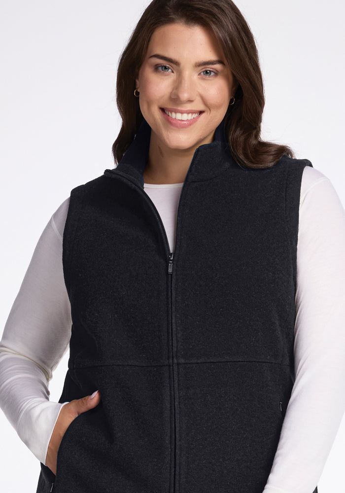 A woman with long brown hair smiles at the camera. She is wearing the Colbie Vest in Carbon Black from Woolx over a white long-sleeved shirt, with her hands resting in its pockets. The background is plain and white.