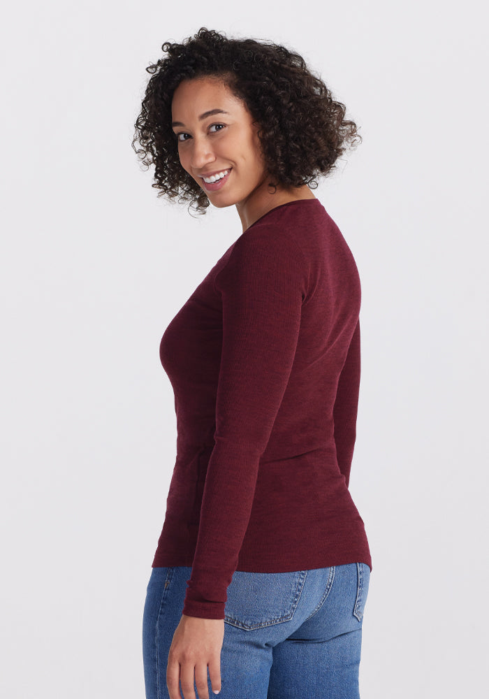 A person with curly hair smiles while posing in the Woolx Reese Ribbed Henley - Cranberry Melange and blue jeans, the Merino wool offering both style and comfort, against a plain background, facing slightly sideways.