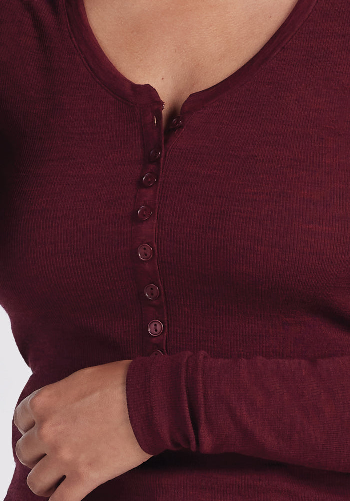 A person is wearing the Woolx Reese Ribbed Henley in a cozy Cranberry Melange, with buttons partly fastened. One arm is crossed over their stomach, slightly obscuring some buttons. The Merino wool fabric appears soft and snug-fitting.