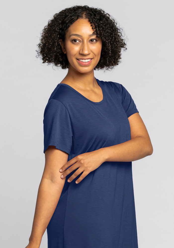 A person with curly hair is smiling in the cozy Desi Nightgown - Starry Night by Woolx. They are posed against a plain light gray background, with one arm bent and the other touching it, exuding a cheerful and relaxed expression.