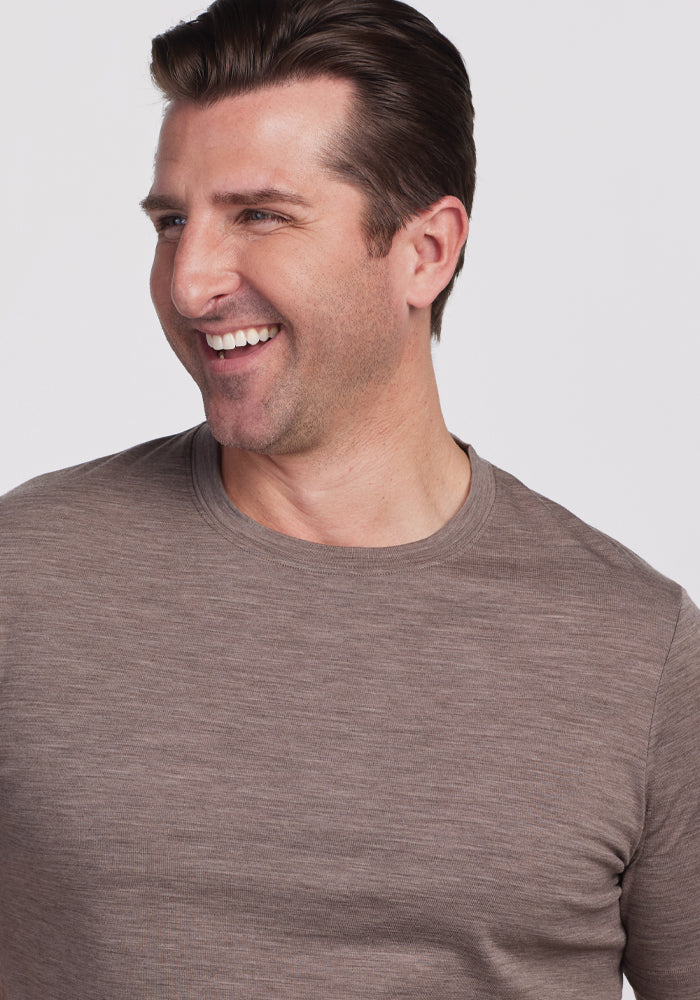 A man is smiling and looking to the side, dressed in a casual Woolx Endurance Tee in Simply Taupe. The background is plain white.