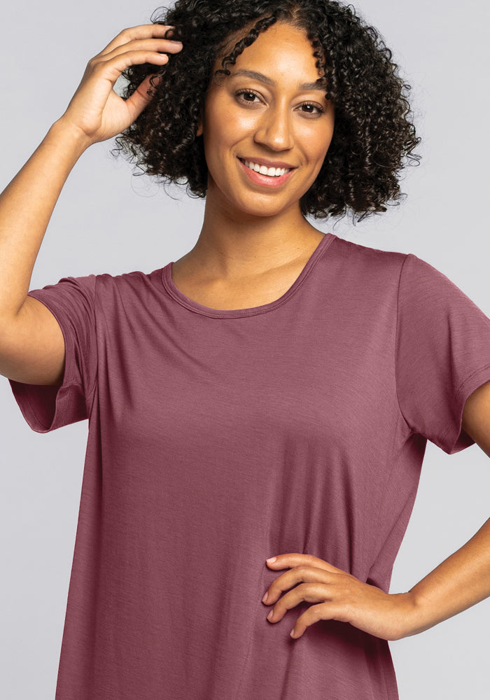 A person with curly hair smiles while posing with one hand on their hip and the other touching their hair. They are wearing the Desi Nightgown - Wild Ginger from Woolx, against a plain gray background.