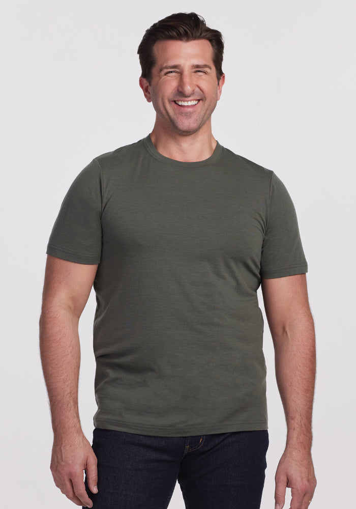 A smiling man in a Woolx Endurance Tee - Deep Fern stands with his hands relaxed by his sides. He has short brown hair and is wearing dark jeans. The background is a plain white. 