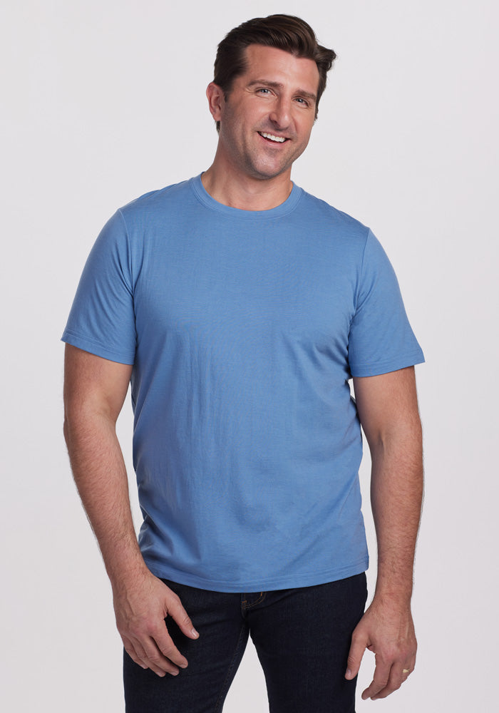 A man with short brown hair stands smiling, wearing a Coronet Blue Endurance Tee by Woolx and dark jeans, showcasing his performance gear against a plain white background. 