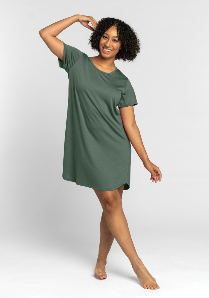 A person with curly hair smiles while posing in a Duck Green Desi Nightgown OLD by Woolx. They stand barefoot against a plain, light gray background, gracefully raising one arm above their head.