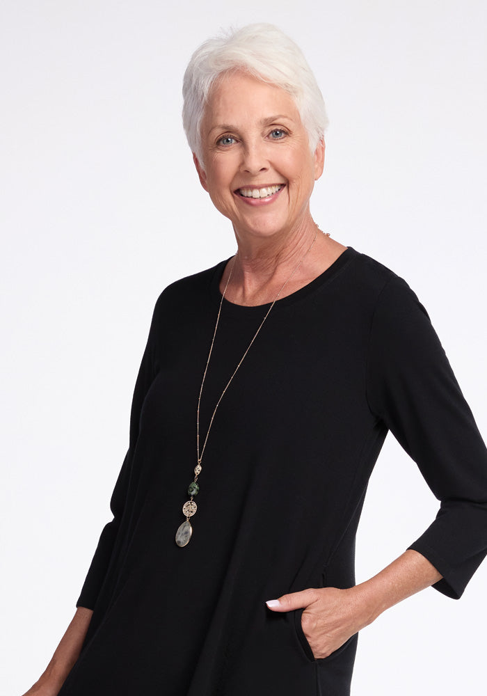 A smiling older woman with short white hair stands against a plain background, wearing the elegant black Rissa A Line Dress by Woolx. Ideal for travel, she complements her outfit with a long necklace featuring decorative pendants, while her left hand is casually tucked in her pocket.