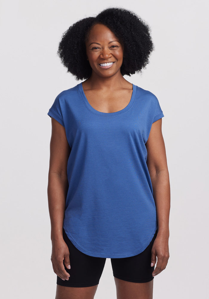 A person with a broad smile stands against a plain background, wearing the blue short-sleeve Woolx Clearance - Gabi Tunic Top made of Australian Merino Wool and black shorts. The person has curly hair and is posed with their arms relaxed by their sides.