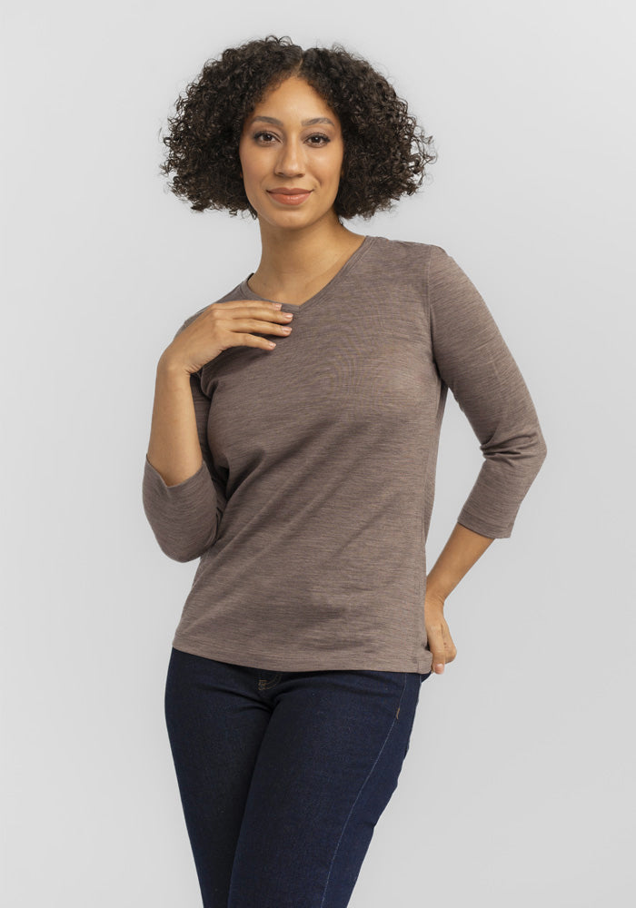 A person with curly hair poses confidently against a plain gray background, wearing a relaxed fit Elena VNeck from Woolx and dark jeans.