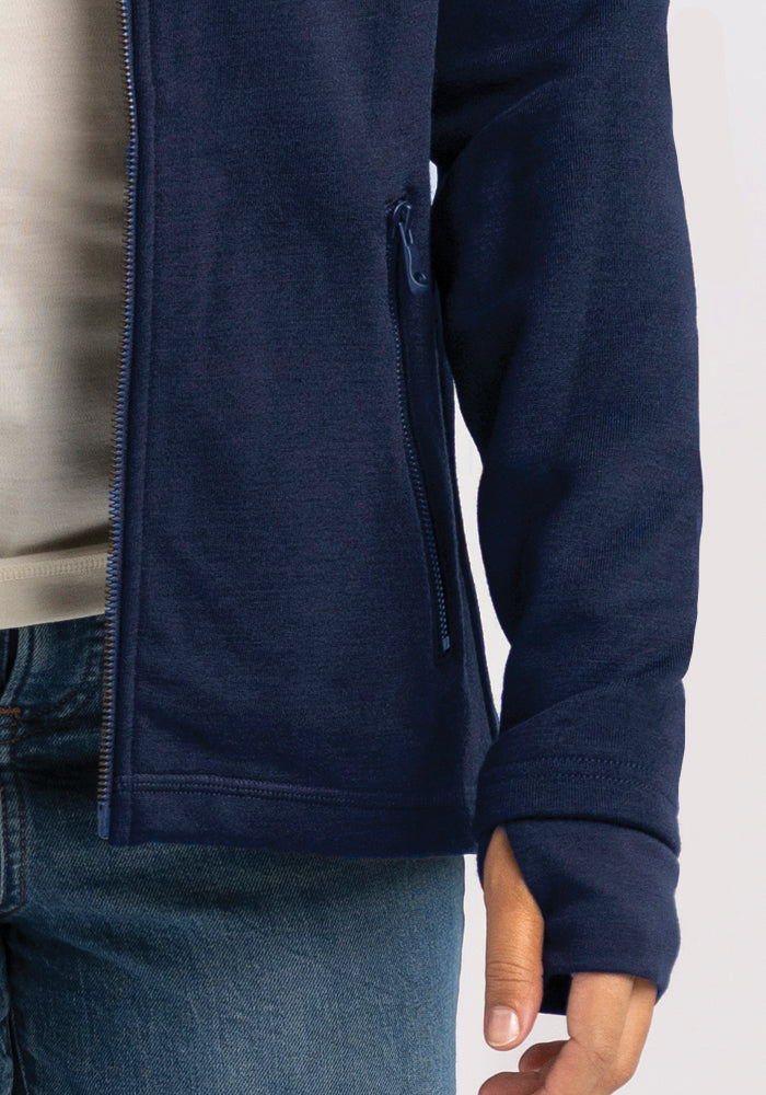 A person is wearing a Woolx Cubby Hooded Sweatshirt in Deep Navy, which combines style with the comfort of the coziest sweatshirt. It features a visible zippered pocket on the right side and thumb holes in the sleeves. Beneath, they have a light-colored shirt and blue jeans. The image focuses on their torso and part of their arms.