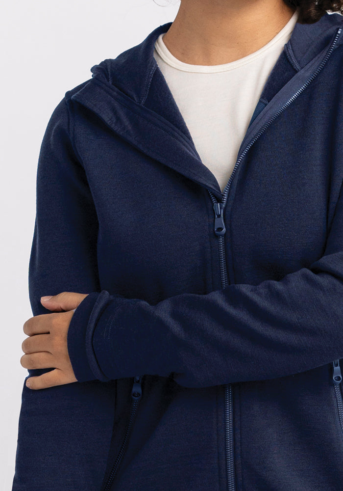 A person in a Woolx Cubby Hooded Sweatshirt - Deep Navy, renowned as the warmest hoodie, crosses their arms over a white t-shirt. The soft texture of the navy blue zip-up stands out against the plain white background, drawing focus to their upper body while keeping their face hidden.