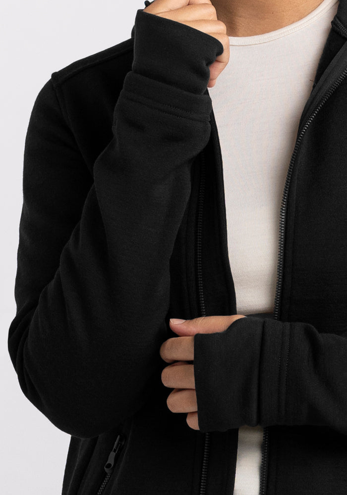 A person wearing a black Woolx Cubby Hooded Sweatshirt over a white shirt is shown from the shoulders to the waist. The person's left hand is partially tucked into their sleeve, with the cuff extending over part of their hand. The background is plain and light-colored.