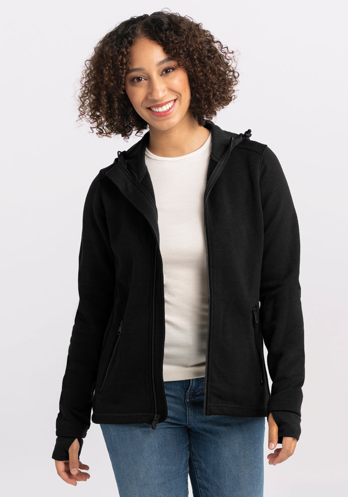 A person with curly hair smiles while wearing a Woolx Cubby Hooded Sweatshirt in Black over a white top and blue jeans. The background is plain and light-colored.