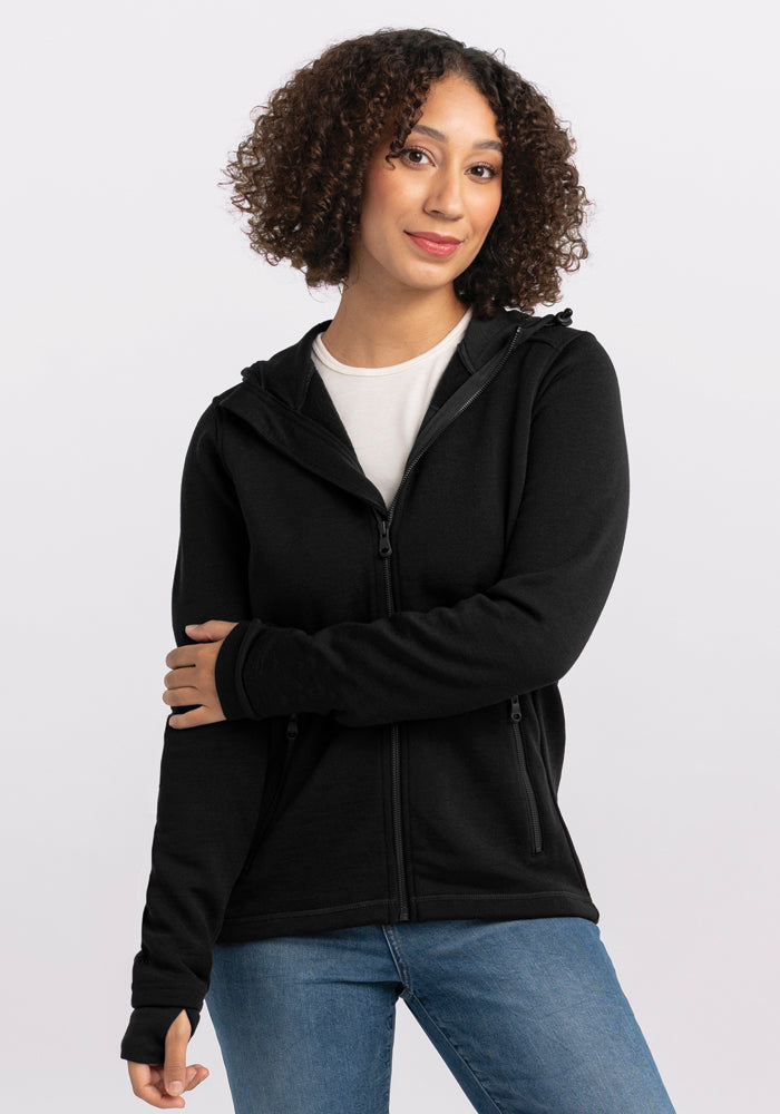 A person with curly hair is wearing a Woolx Cubby Hooded Sweatshirt in black with heavy-duty zippers over a white shirt and blue jeans. They are standing against a plain light-colored background, with one arm crossing their body and the other hanging by their side. 