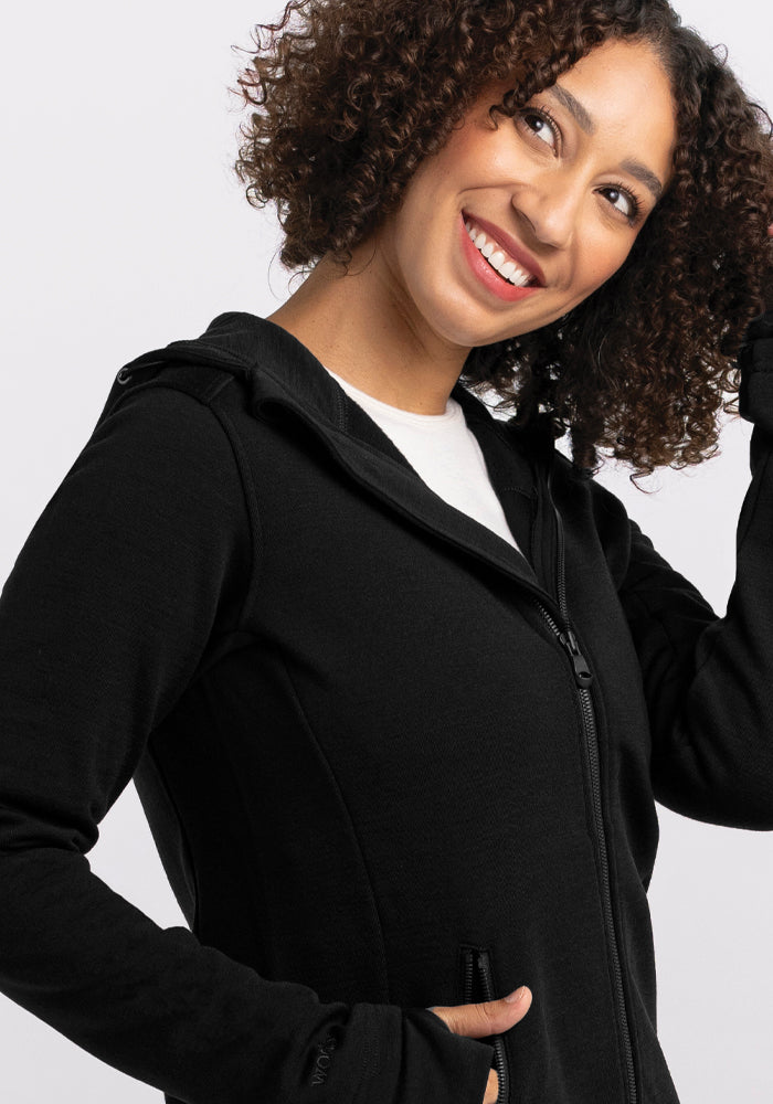 A smiling woman with curly hair poses against a plain background. She is wearing a Woolx Cubby Hooded Sweatshirt in Black with a white shirt underneath. One of her hands is tucked in a pocket, while the other touches her hair.