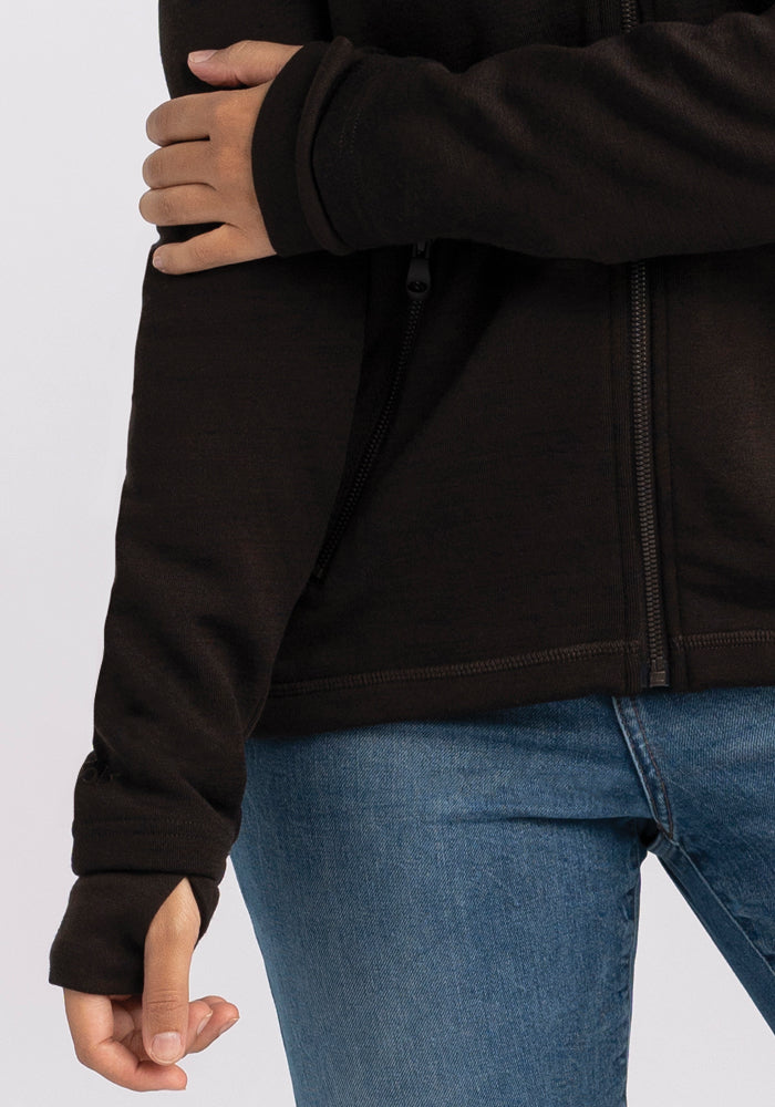 A person wearing a Woolx Cubby Hooded Sweatshirt in French Roast and blue jeans is shown from the shoulders to the hips. Their left arm is bent, with the right hand resting over the left arm at elbow level. The sweatshirt features a thumb hole in the sleeve for added comfort.