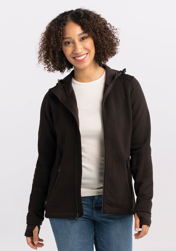 A woman with curly hair is smiling at the camera. She is wearing a white shirt under a Woolx Cubby Hooded Sweatshirt in French Roast and blue jeans. The background is plain white. 