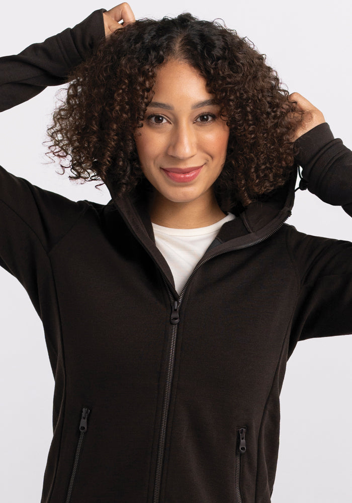 A person with curly hair smiles while adjusting the hood of their Woolx Cubby Hooded Sweatshirt in French Roast, which is partially zipped over a white shirt. The background is a plain, light gray.