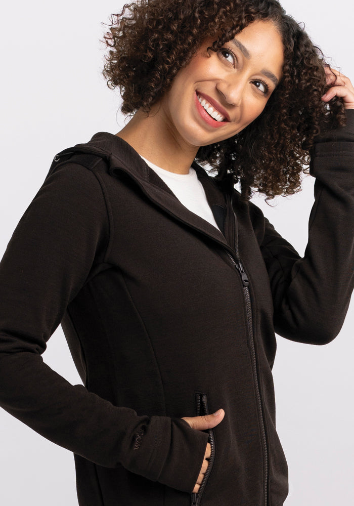 A person with curly hair smiles while wearing the cozy Woolx Cubby Hooded Sweatshirt in French Roast. They are looking upwards and touching their hair with one hand, against a plain, light gray background.