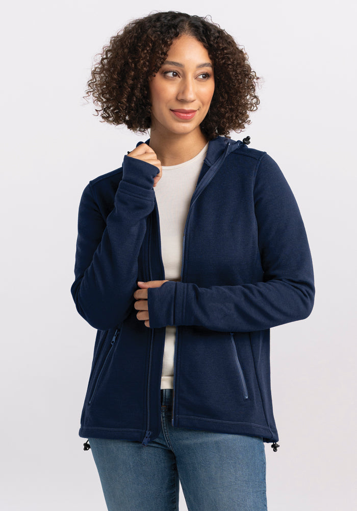 A person with curly hair is wearing a deep navy Cubby Hooded Sweatshirt from Woolx over a white shirt. They are looking slightly to the side and have one hand near their chin. The hoodie features zipped pockets and a drawstring at the waist. The background is plain and light-colored. 