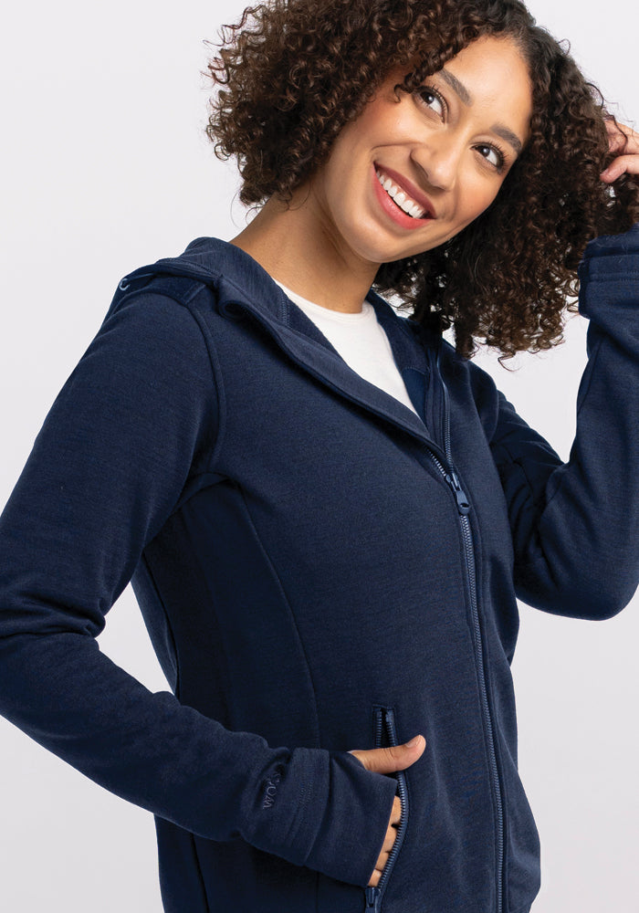 A woman with curly hair smiles and gazes to the side, wearing a Woolx Cubby Hooded Sweatshirt in Deep Navy. Her left hand gently touches her hair against a plain white background, emphasizing the cozy warmth of this snug sweatshirt.