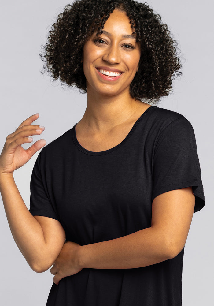 A person with curly hair smiles at the camera, wearing a Desi Nightgown in black by Woolx. One hand is raised near their shoulder against a neutral background, exuding the cozy vibe of comfort sleepwear.