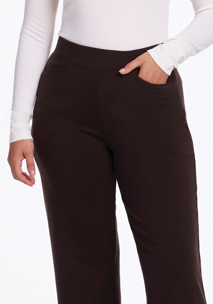 A person dressed in a white long-sleeve top and Woolx's Ellie Wide Leg Pants in French Roast, with one hand casually tucked into the side pocket. The plain, light-colored background highlights the refined merino wool ensemble effortlessly.