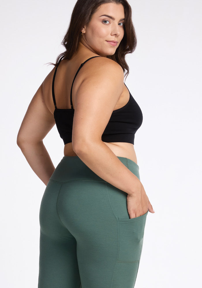 A person with long, dark hair stands sideways and glances over their shoulder at the camera. They are dressed in a black crop top and high-waisted Piper Pocket Leggings in Duck Green by Woolx, with their hand resting in the side pocket. The background is white.