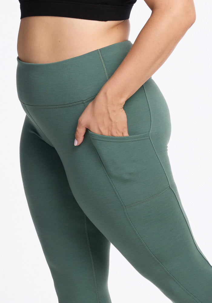 A person wearing Woolx's Piper Pocket Leggings in Duck Green and a black sports bra has their right hand placed inside the side pocket, emphasizing the functionality of the leggings' non-see-through merino wool fabric. The image spotlights their lower torso and thighs against a plain background.