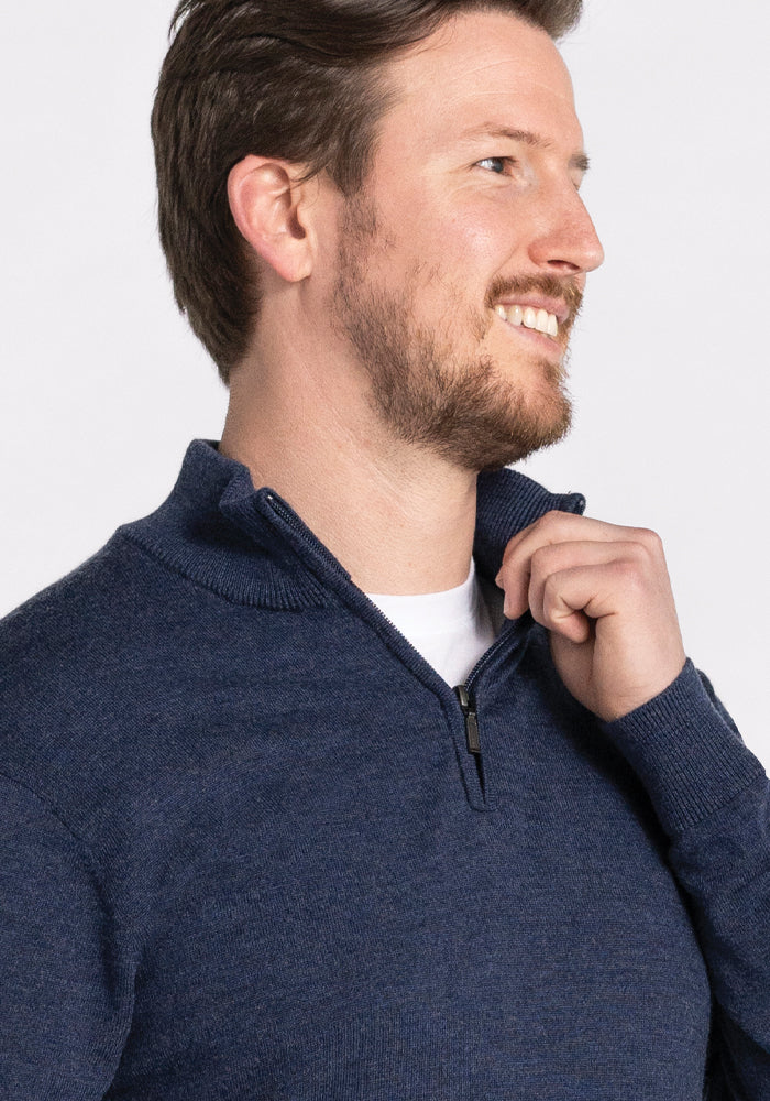 A man with a beard smiles while looking to the side, dressed in the Woolx Enzo Quarter Zip - Twilight Storm over a white shirt. He holds the zipper pull near his chin against a light-colored background.