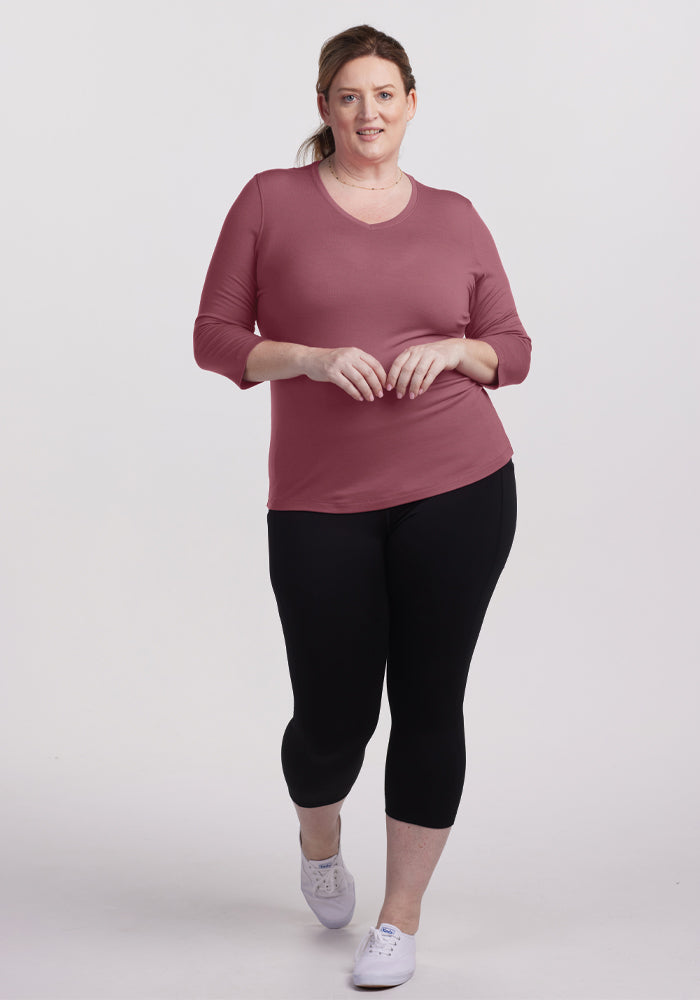 A woman wearing a Woolx Elena V-Neck Top in Wild Ginger, black leggings, and white sneakers strides confidently against a plain white background.