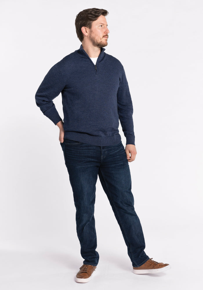 A bearded man stands against a white background, wearing the Enzo Quarter Zip in Twilight Storm by Woolx. He pairs it with dark jeans and brown shoes, with his right hand casually in his pocket as he gazes to his left.